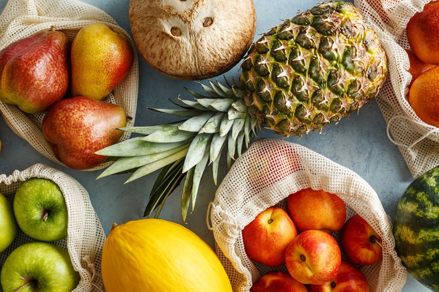Verschiedene Früchte in wiederverwendbaren Netzbeuteln: Ananas, Kokosnuss, Birnen, grüne Äpfel, Nektarinen/Äpfel sowie gelbe und Wassermelone auf heller Oberfläche.