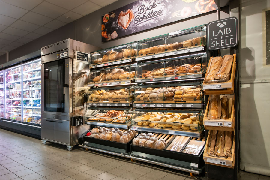 Backwarenregal mit Broten, Brötchen und Baguettes neben Ofen im Markt.