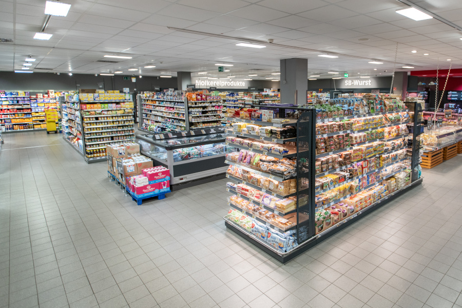 Supermarktgang mit Regal voller Brotsorten und Backwaren; im Hintergrund Kühltruhen und Beschilderung „Molkereiprodukte“ und „SB-Wurst“.