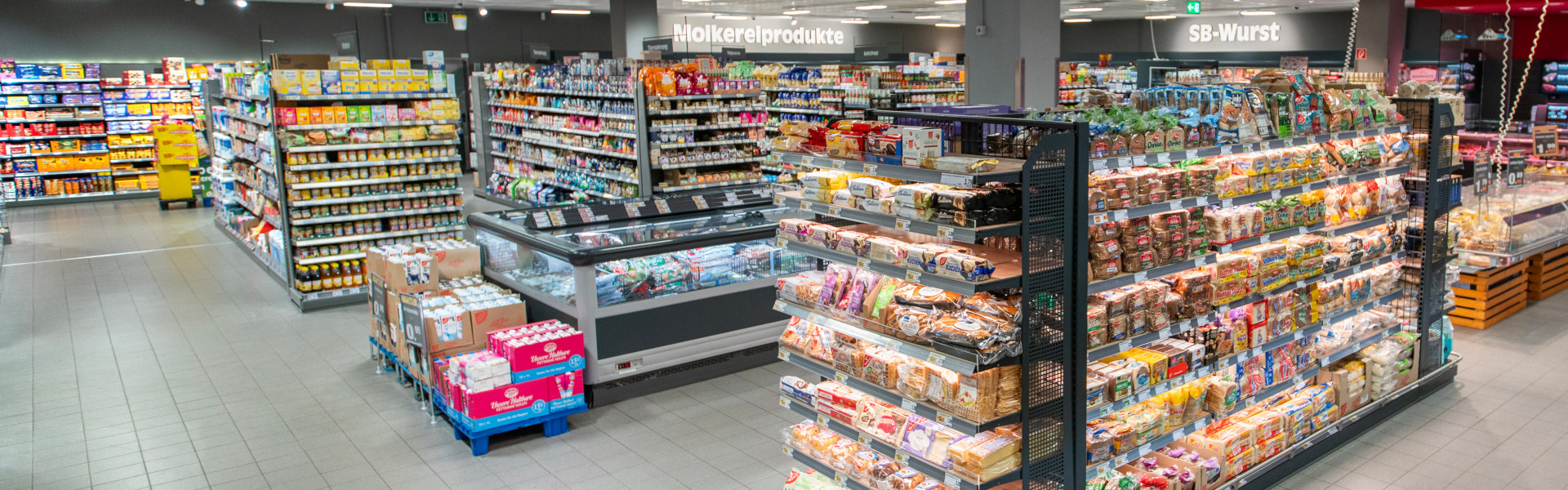 Supermarktgang mit Regal voller Brotsorten und Backwaren; im Hintergrund Kühltruhen und Beschilderung „Molkereiprodukte“ und „SB-Wurst“.