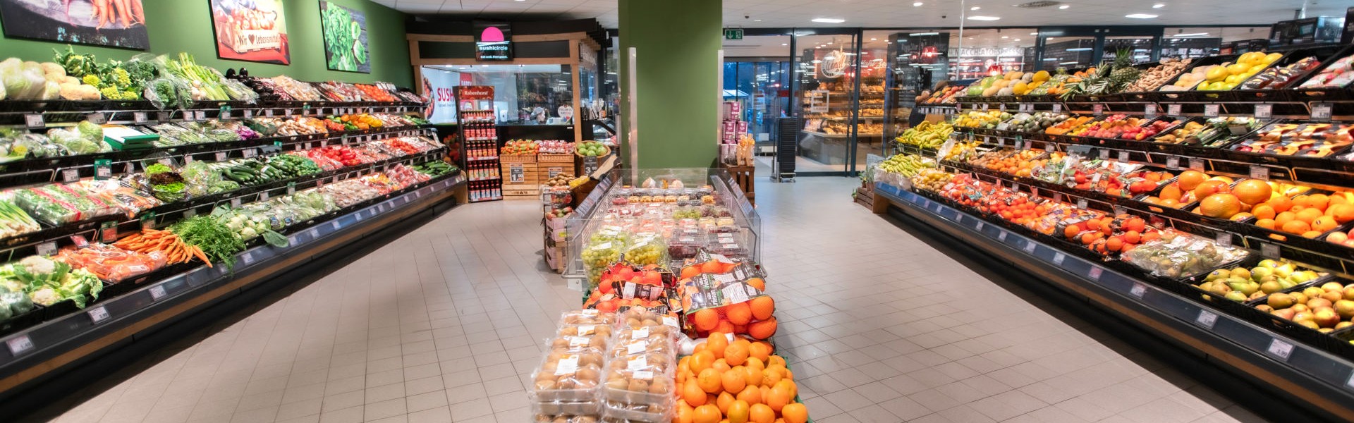 Große Obst- und Gemüseabteilung mit beidseitigen Regalen; mittig Auslage mit Orangen und Kartoffeln, hinten Blick zur Bäckerei.