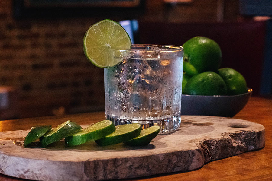 Gin Tonic im Tumbler mit Limettenscheibe; Limettenspalten auf Holzbrett, Schale mit Limetten im Hintergrund.