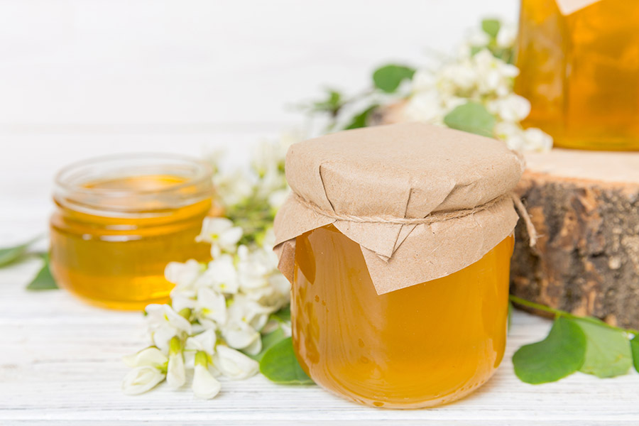 Honigglas mit braunem Papierdeckel, daneben weiteres Glas und weiße Blüten im Hintergrund.