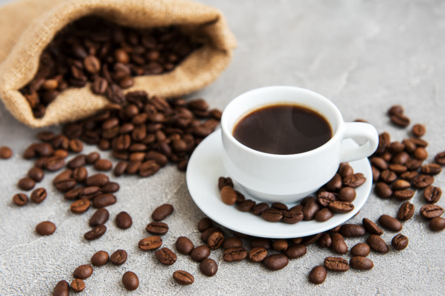 Weiße Tasse mit schwarzem Kaffee auf Unterteller, umgeben von verstreuten Kaffeebohnen und einem offenen Jutesack.