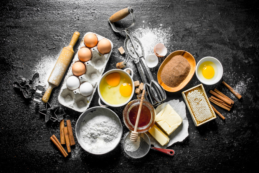 Backzutaten auf dunkler Fläche: Eier, Mehl, Butter, Zucker, Zimt, Honig, Nudelholz, Schneebesen und Ausstechformen.
