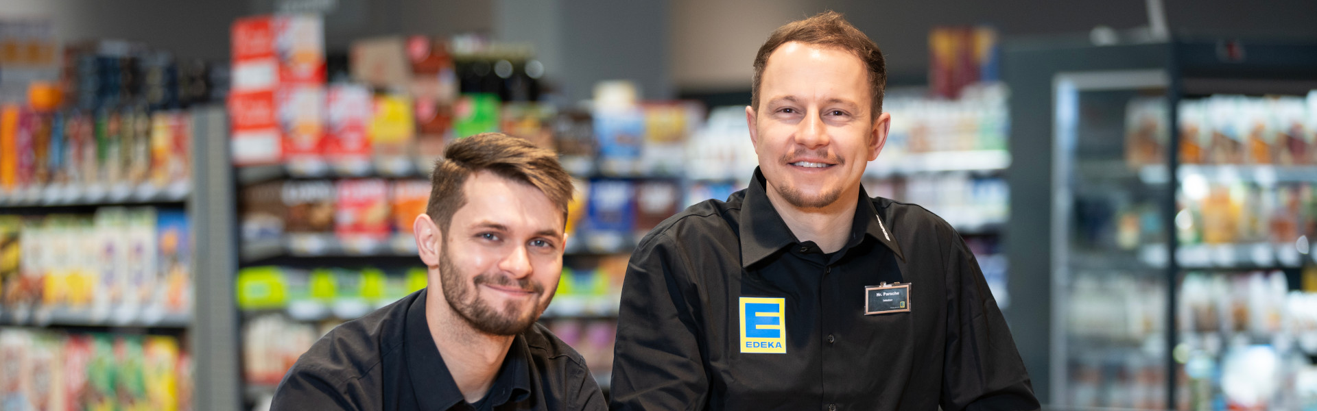 Zwei EDEKA-Mitarbeiter in schwarzer Dienstkleidung lächeln in die Kamera; unscharfe Marktregale im Hintergrund.