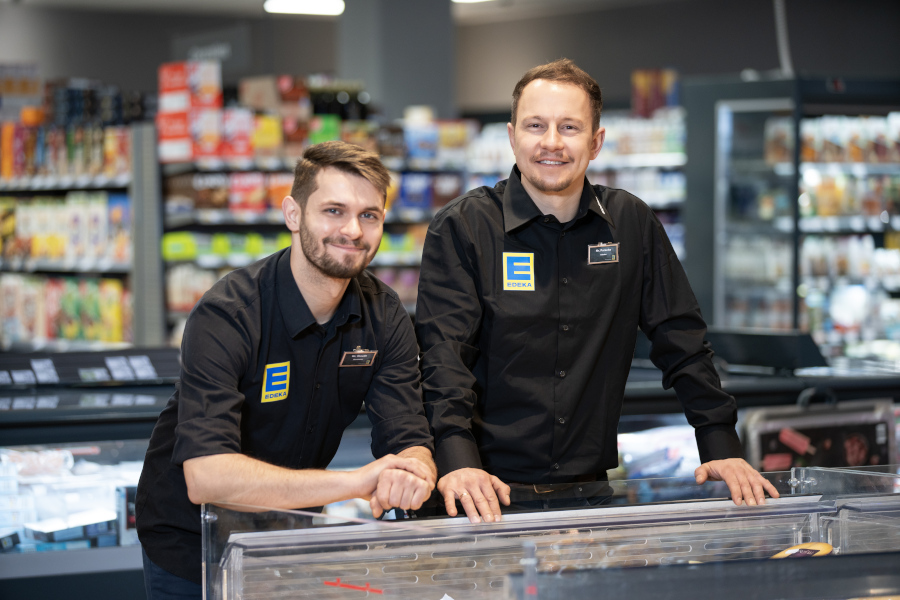 Zwei EDEKA-Mitarbeiter in schwarzer Dienstkleidung lächeln in die Kamera; unscharfe Marktregale im Hintergrund.